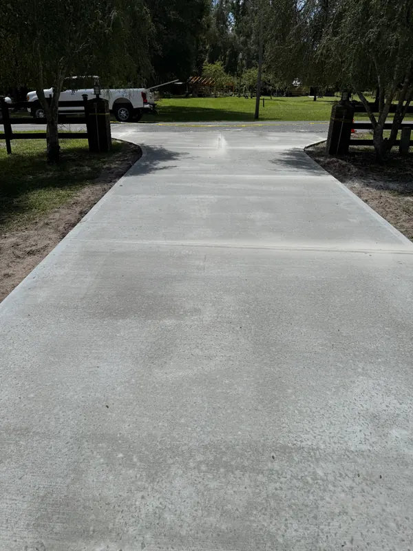 Straight broom-finish concrete driveway running between wooden fence posts under willow trees, with an even brushed texture and clean control joints