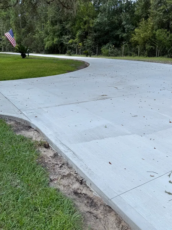 Wide broom-finish concrete driveway apron curving out to meet a rural road, with tooled edges, a lawn on the left and an American flag by the treeline