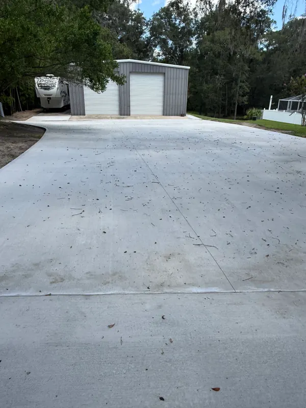 Wide broom-finish concrete pad running up to a gray metal shop building with a roll-up door, with a fifth-wheel RV parked alongside it