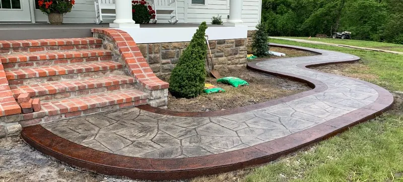 Curved stamped concrete walkway in a random flagstone pattern with a deep red-brown decorative border, sweeping past brick front steps and a stone foundation