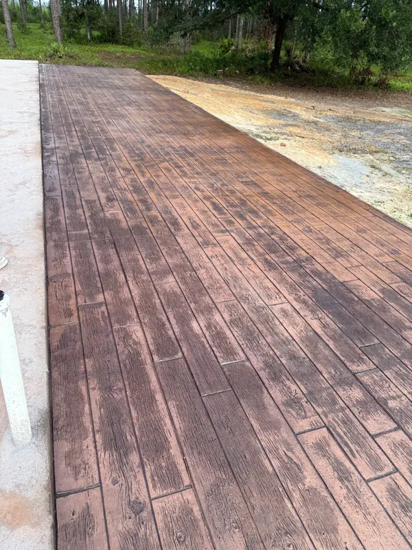 Wood-plank stamped concrete slab photographed damp, deepening the walnut and espresso tones, with a Florida pine treeline behind it and fresh sand fill along the edge